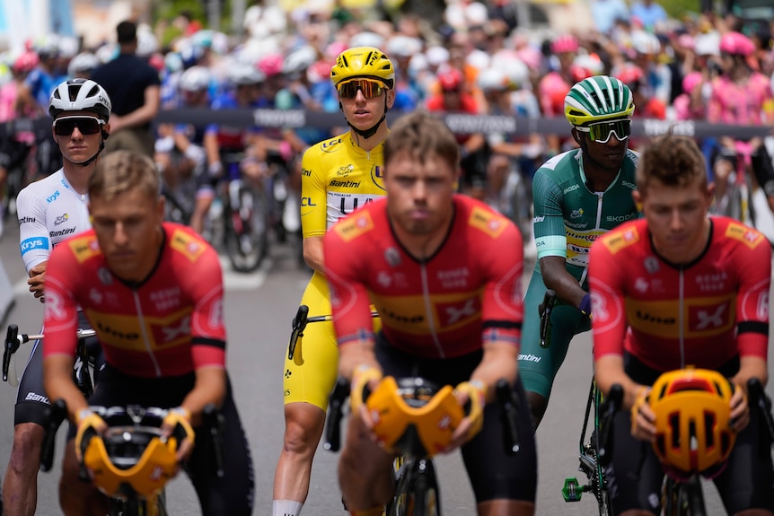 A group of cyclists in red stare silently at the camera, with the race leaders lined up behind them.