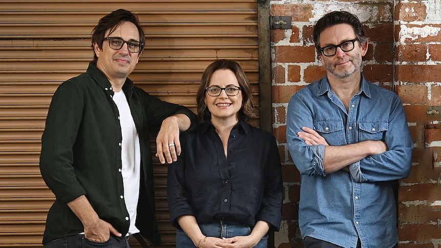 Trent Dalton, Fiona Franzmann, Sam Strong standing together against an orange roller door and brown brick wall