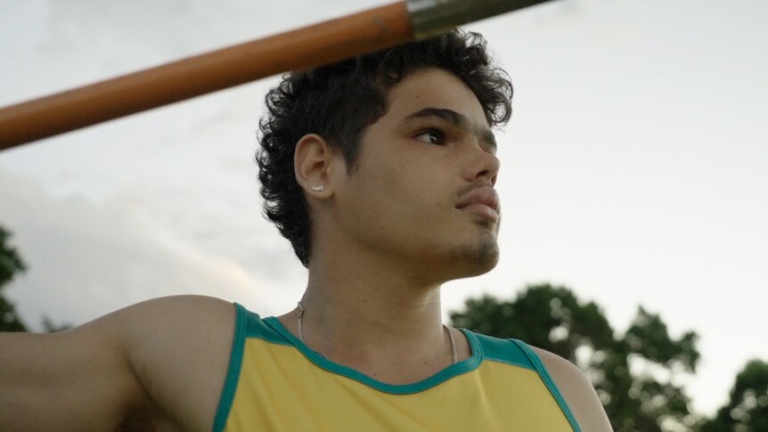 A young Indigenous teenager in a yellow and gold shirt holding a javelin. 