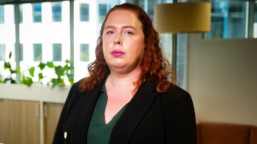 A young woman with red hair in corporate attire looks sternly at the camera.