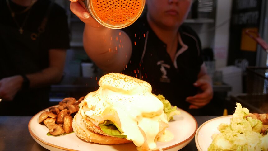 A young woman sprinkles paprika on a serving of avocado on toast with a poached egg