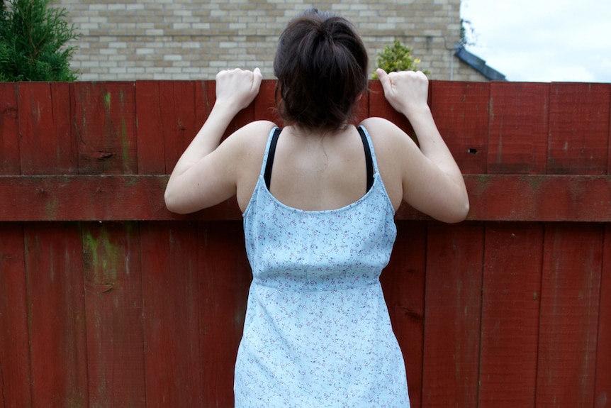 A woman with brown hair wearing a white sundress peeps over a fence.