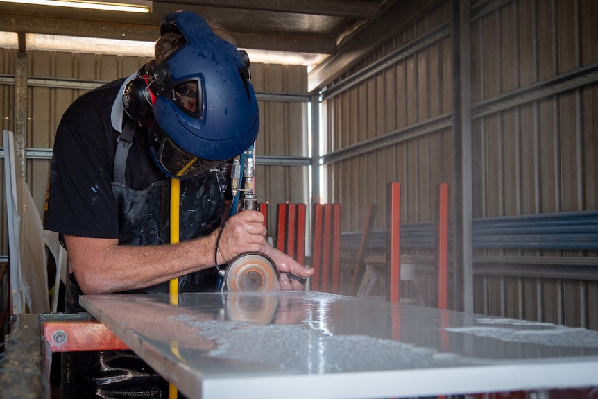 A man at a workbench cuts a piece of engineered stone. He wears a full head covering.