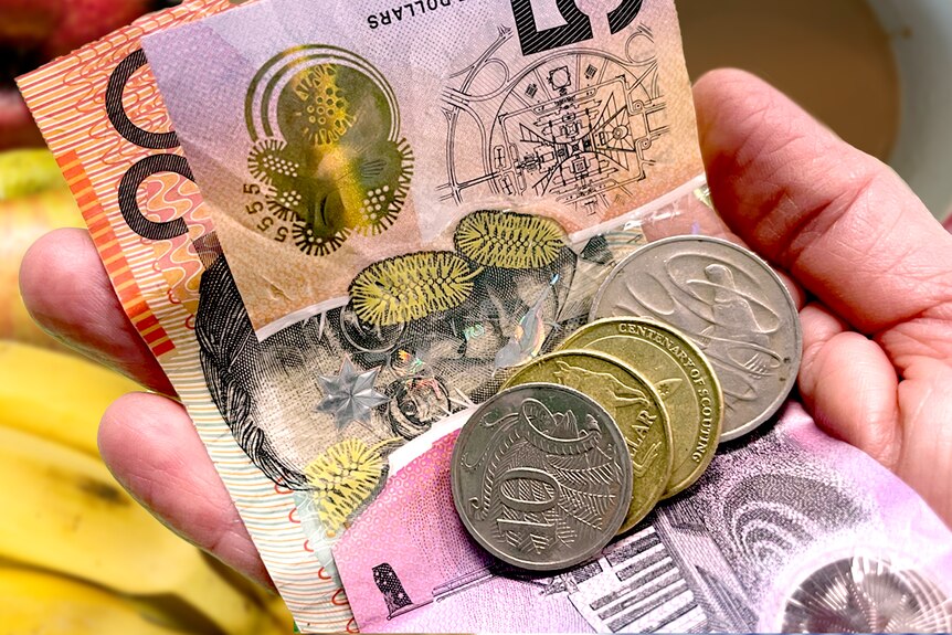 Hand holding money, notes and coins with a coffee cup and fruit in the background, cost of living