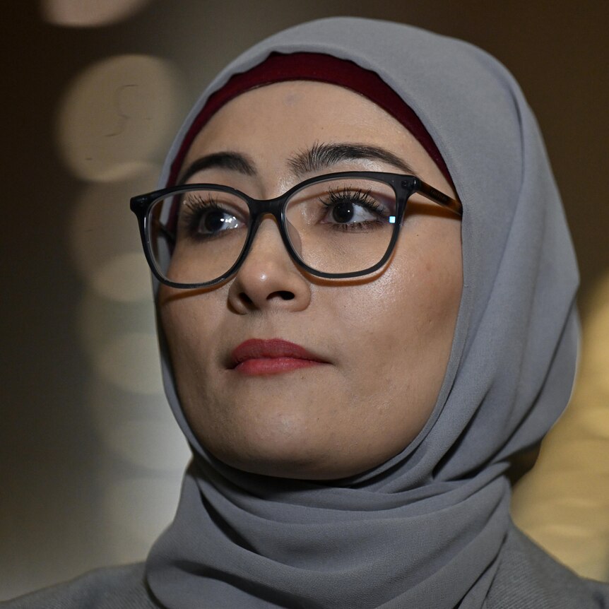 A young woman wearing thin glasses, light red lipstick and a grey hijab looks to her right in closeup