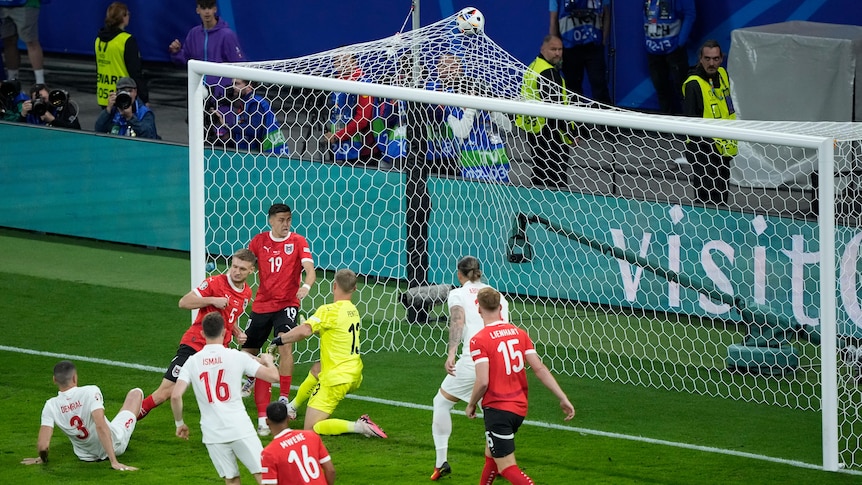 A group of Türkiye and Austria footballers stand around, as the ball hits the top of the net - the goalscorer sits on the turf.