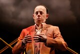 A man in his 50s in an orange suit stands on a stage, cloud and golden abstract structure in the b/g, his hands reaching togeth