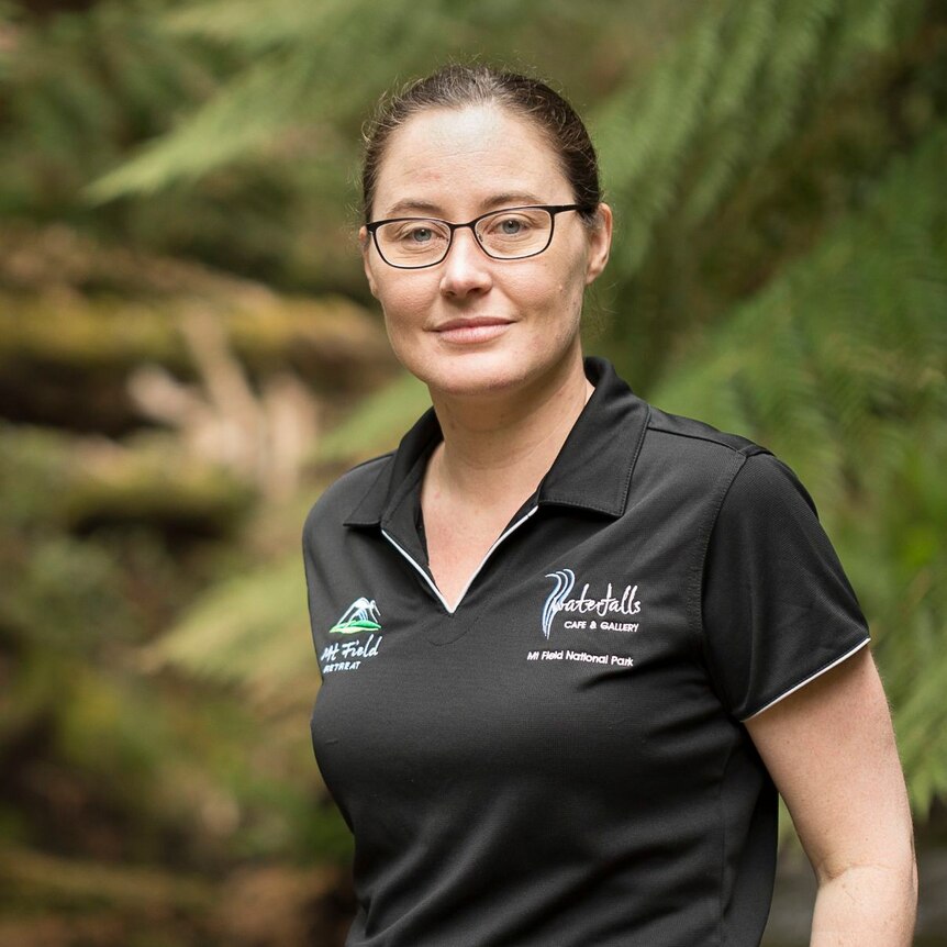 A young woman wearing spectacles looks at the camera while standing outside with ferns behind her.