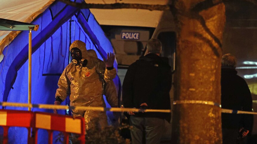 Investigators in chemical hazmat suits walk next to a police tent.