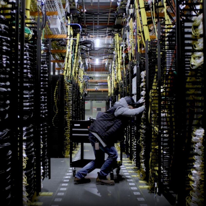 A man leans to work on a large server, standing in an aisle of servers with cables running everywhere.