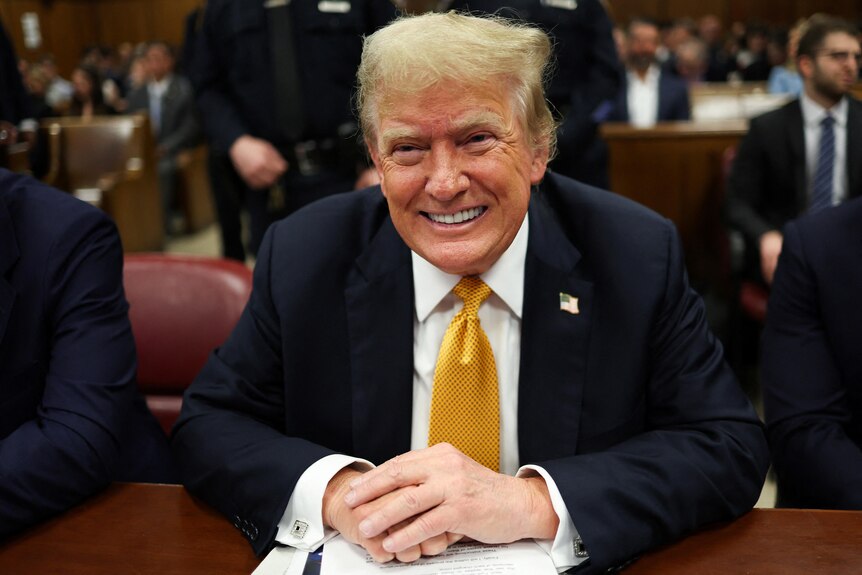 Trump smiles in a yellow tie and suit in a court