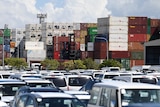 Rows of cars in a car park, with stacked shipping containers behind them.