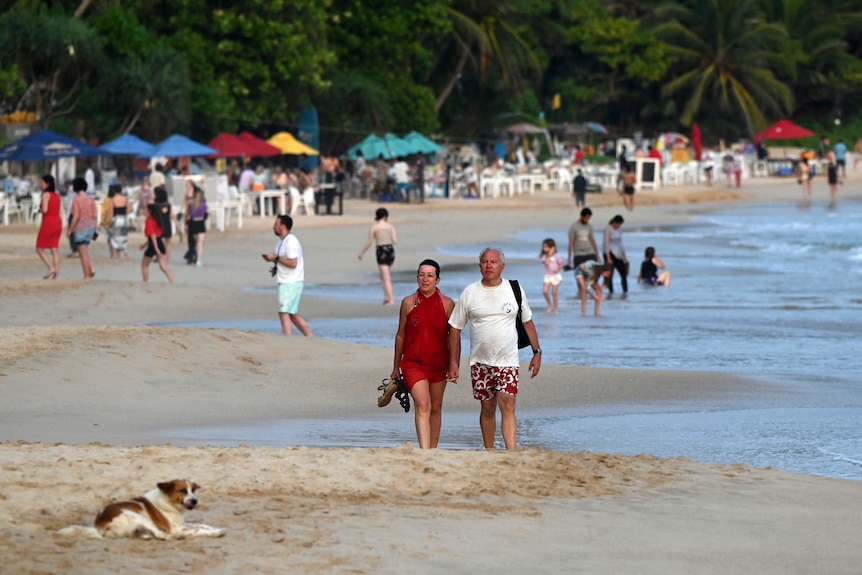 A man and a woman walk along a yellow sand beach and colourful umbrellas dot the shore.