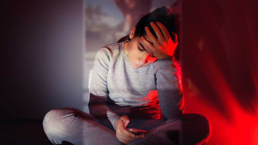 A girl sitting cross-legged looking at her phone with a worried look on her face; and her forehead in the palm of her hand