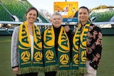 Three women wearing green and yellow Australia scarves stand in an empty stadium to pose for a photo