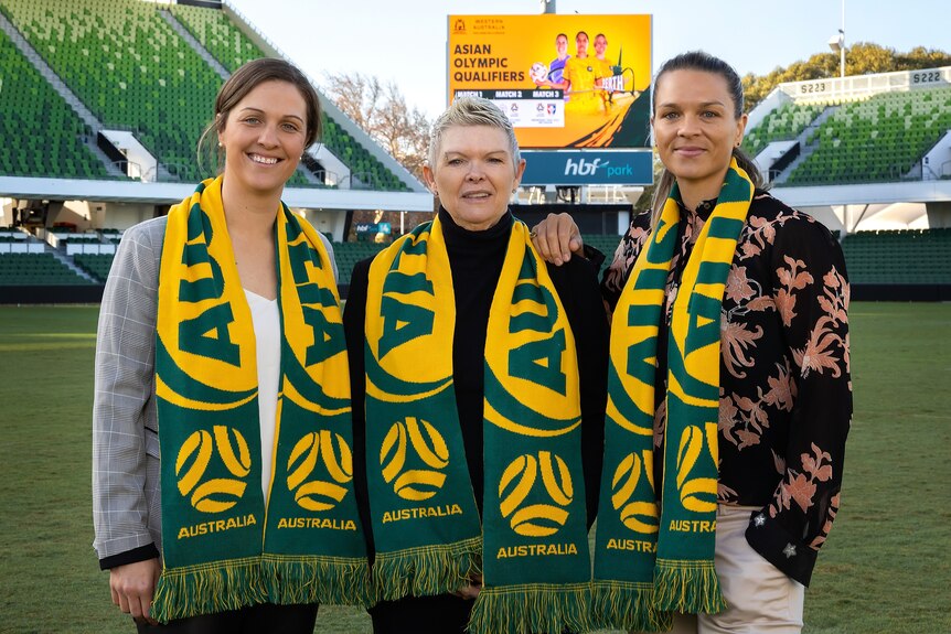 Three women wearing green and yellow Australia scarves stand in an empty stadium to pose for a photo