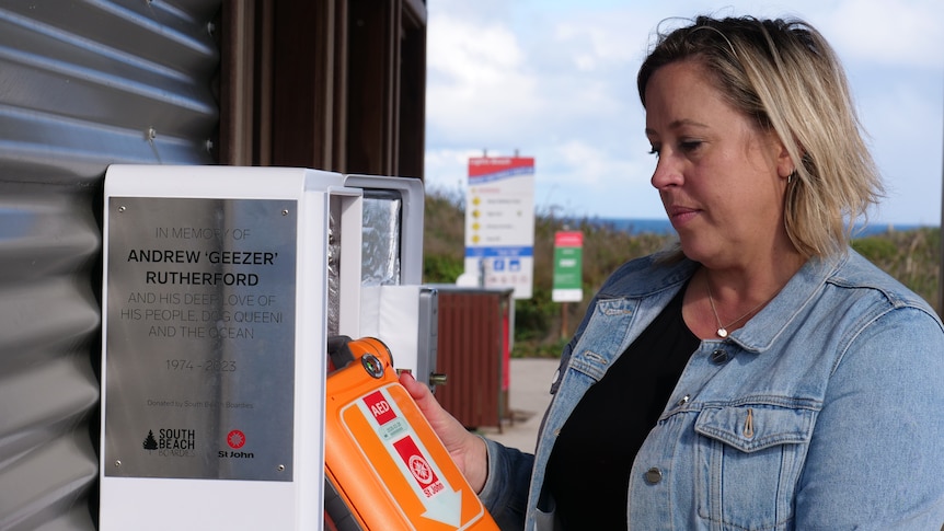 Renee holding a defibrillator next to its box. The box has a plaque to Andrew 'Geezer' Rutherford.