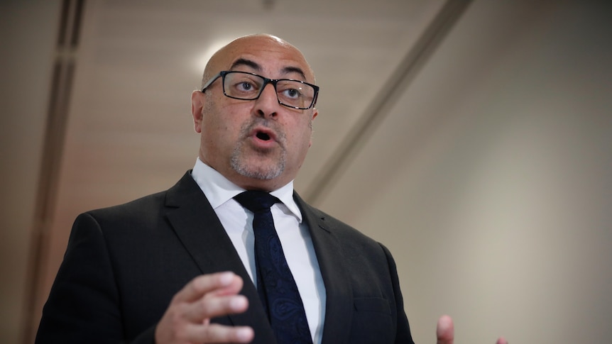 A man in a suit and tie and glasses speaks and gestures with his hands.