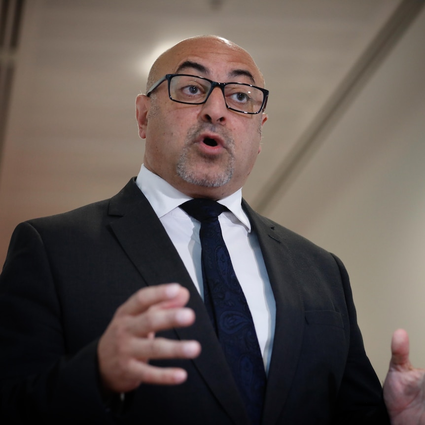 A man in a suit and tie and glasses speaks and gestures with his hands.