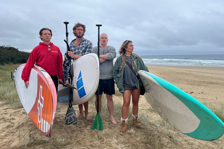 Three men and a woman holding paddle boards and paddles stand on a sand dune looking out at the ocean.