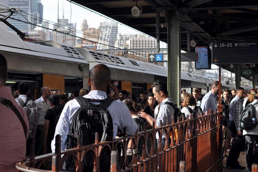 A crowded outdoor train station with people getting off and on of trains. Others talk and look at phones.