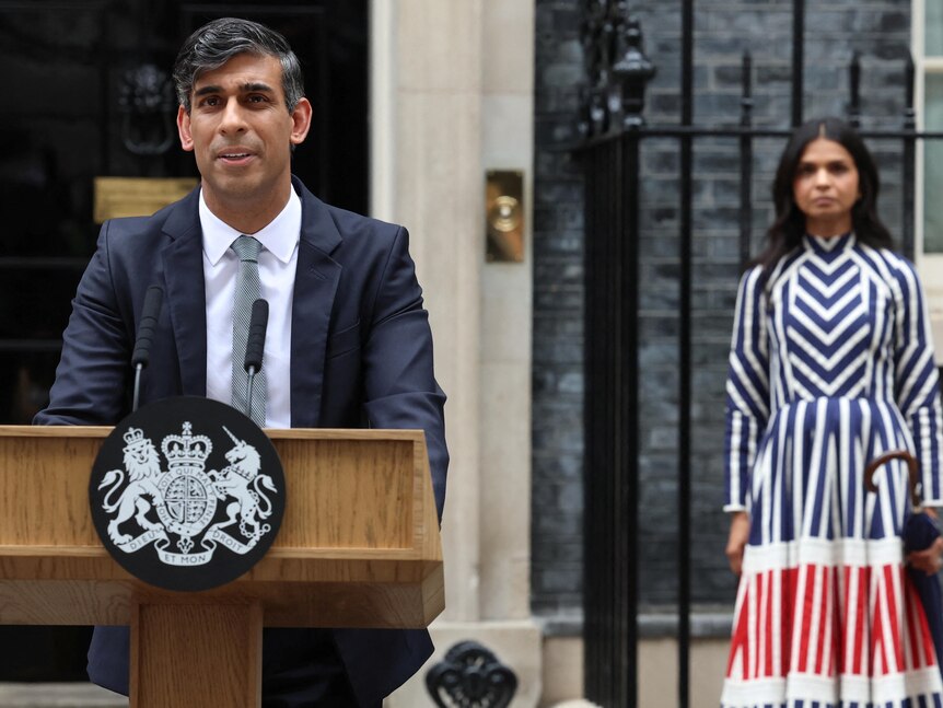 Rishi Sunak at a lecture his wife stands in a striped dress in the background. 