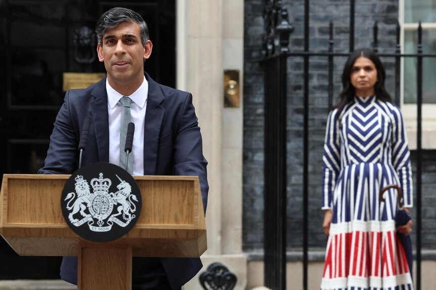 Rishi Sunak at a lecture his wife stands in a striped dress in the background. 