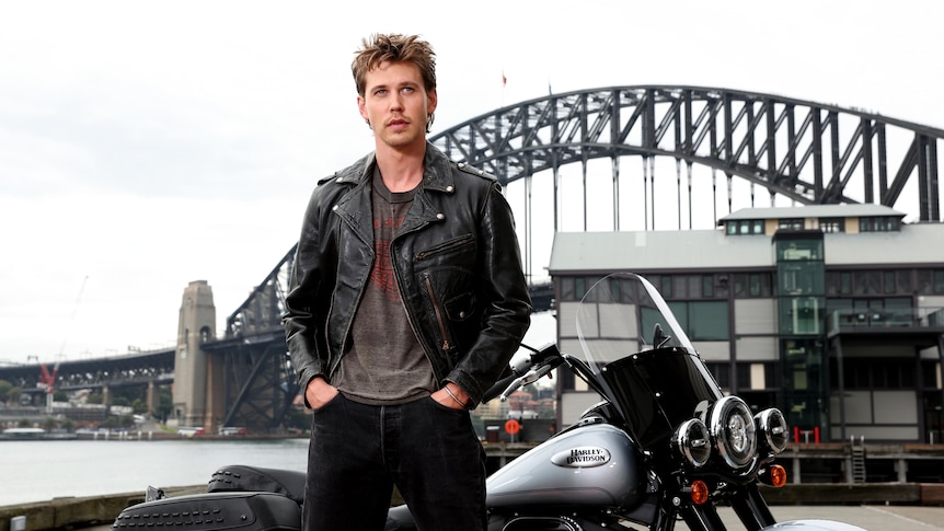 Austin Butler in a leather jacket stands by a motorbike, with the Sydney Harbor Bridge behind him