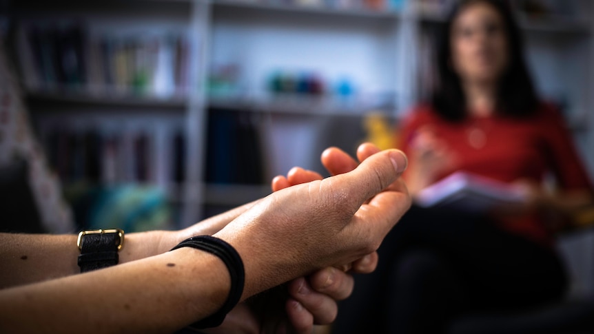 A close up of fidgeting hands. In the background, a psychologist watches on, taking notes.