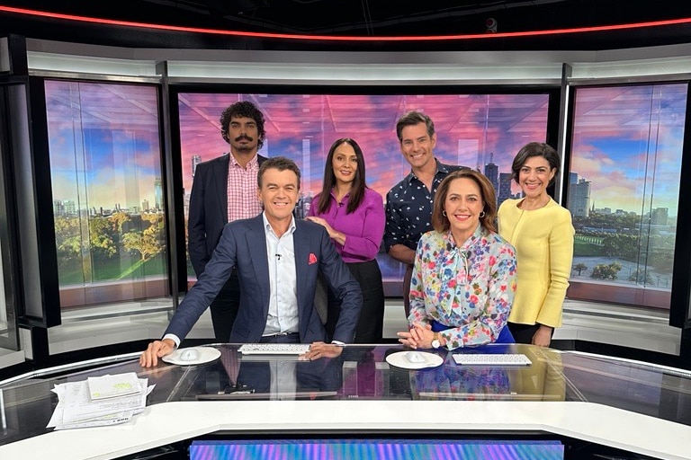 Three women and three men pose for a photo around a TV studio desk.