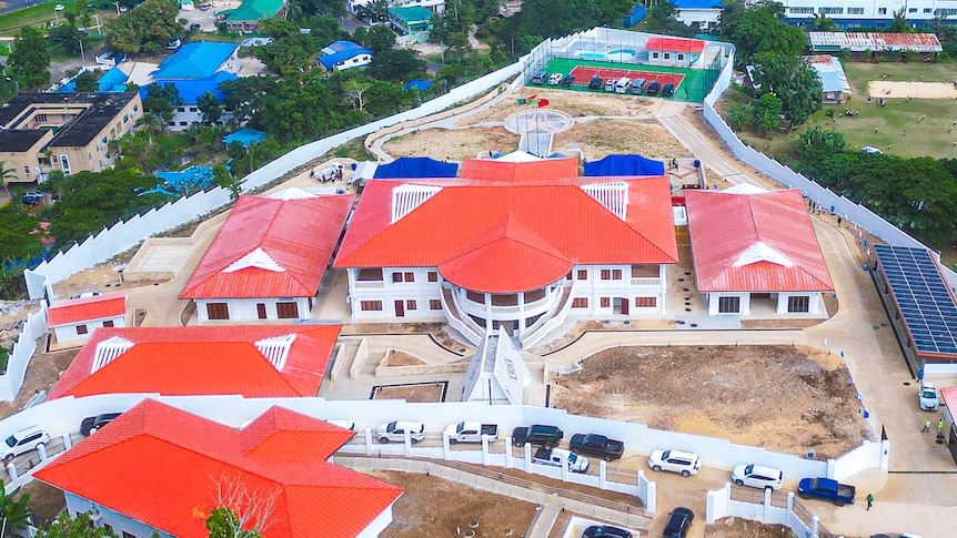 An aerial shot shows a large complex of buildings with red roofs, including a palace, with a bay in the background.