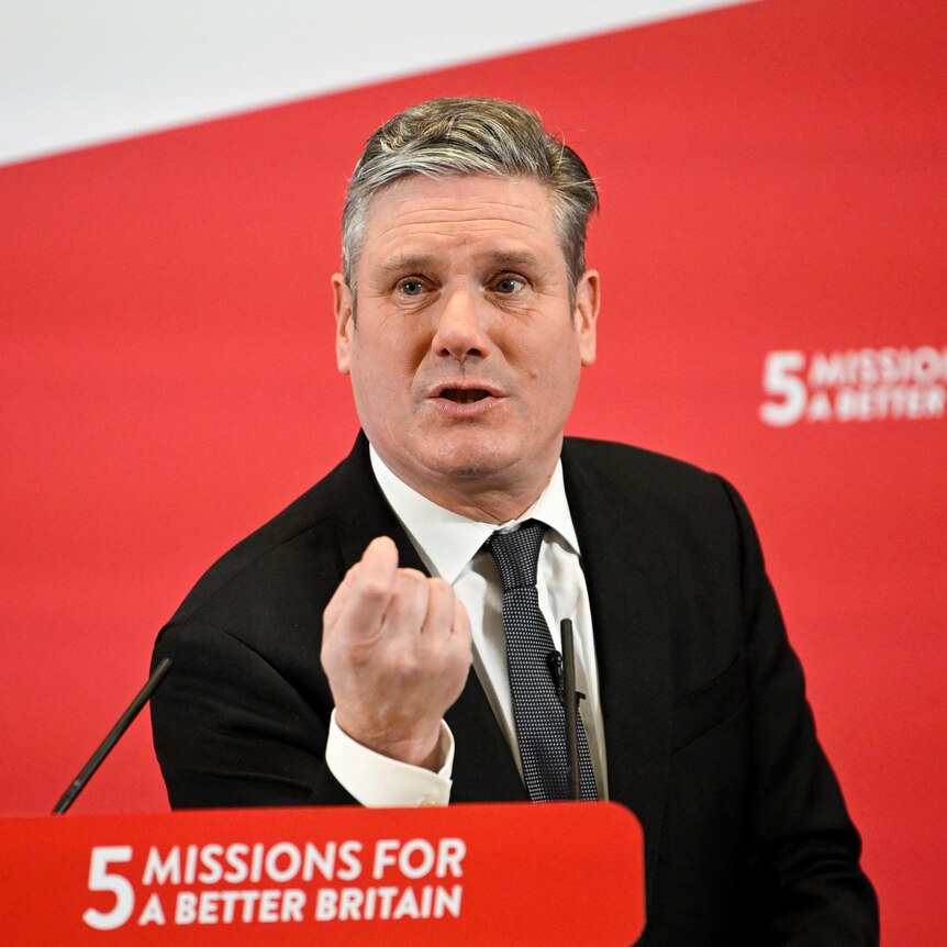 A man stands behind a lectern inside in front of a red banner that says '5 missions for a better future'