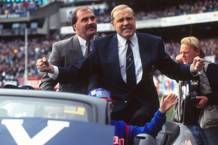 An emotional man in a suit and tie with a 'V' on it stands in a car with fists clenched as he does a farewell lap of the MCG.