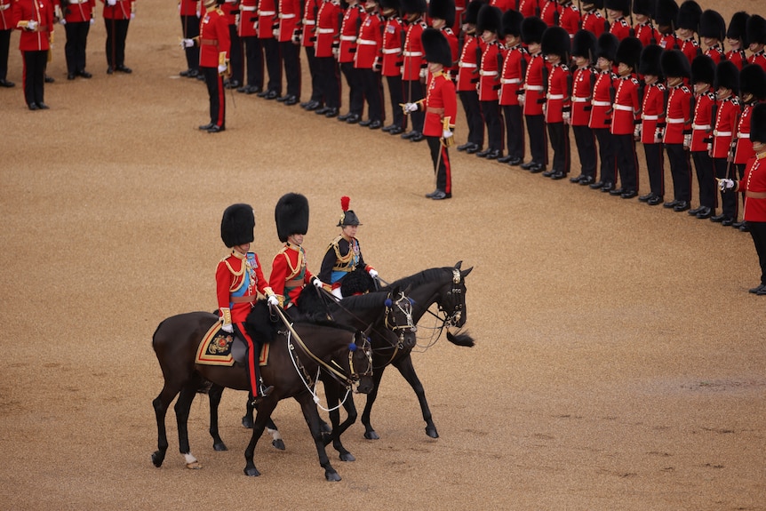 Three riders atop dark-coloured horses ride in military uniform past a line of soldiers