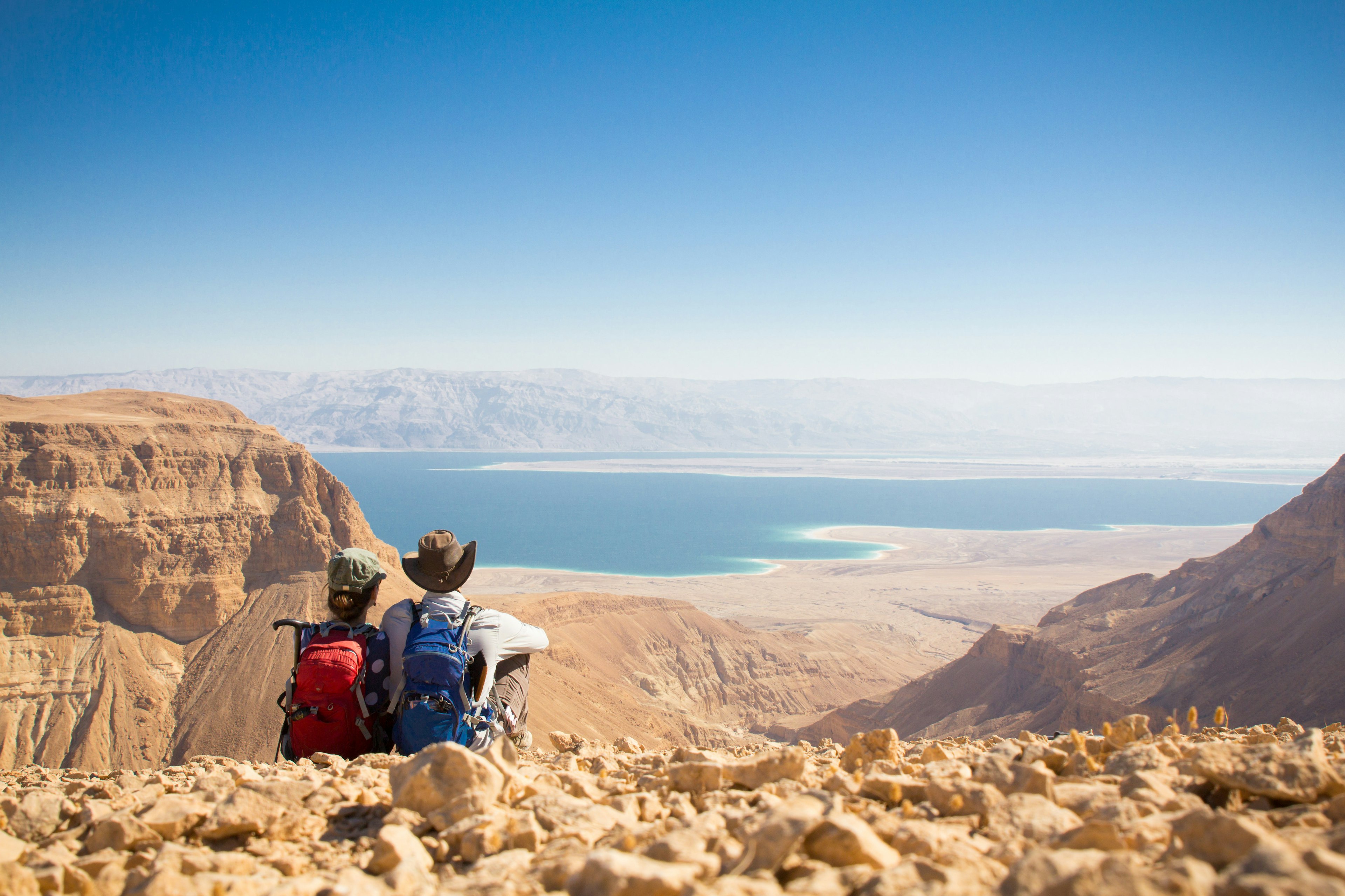 A hiking couple sitting on the edge of a mountain overlooking a large body of water.