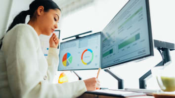 A woman in front of two monitors looks at data reports and writes in a notebook. 