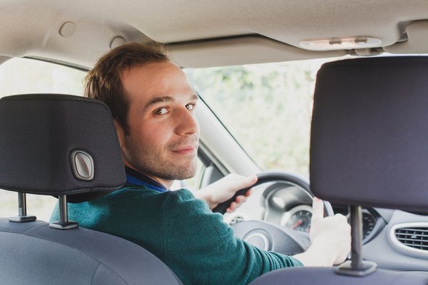 We see a young man in a car's driver's seat, turning around to look at someone in the back seat.