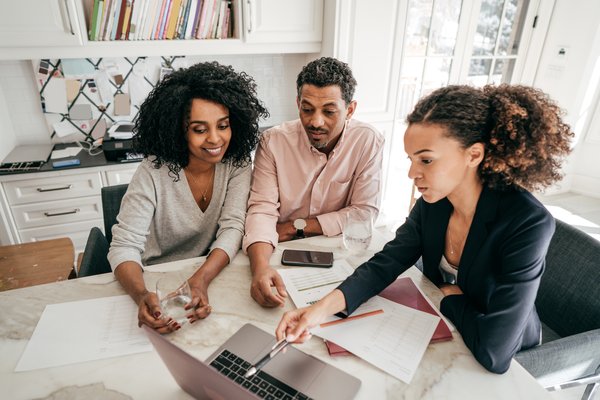 Three people sitting at a document-covered table in a home office and discuss information on a laptop screen.