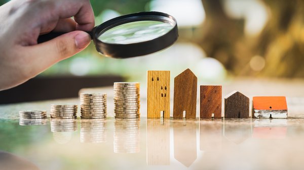 A person holding a magnifying glass looking at a row of rising coins and buildings.