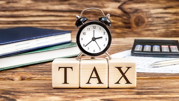 An alarm clock resting atop little wooden blocks that spell out TAX and sitting on a desk with books and a calculator.