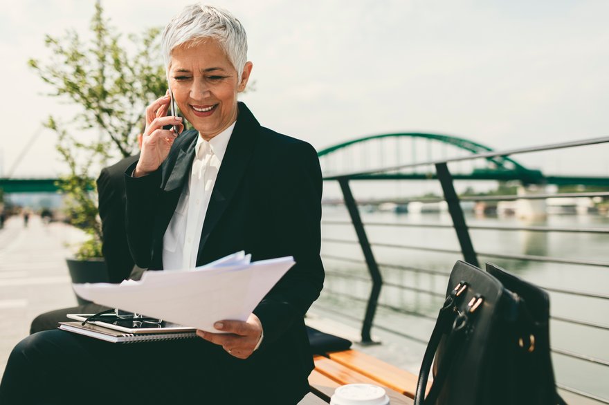 Person sitting on a bench near a waterway and bridge while talking on the phone and looking at documents.