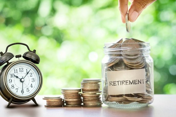 Hand placing a coin into a glass jar labeled "retirement" with stacked coins and an alarm clock next to it.  