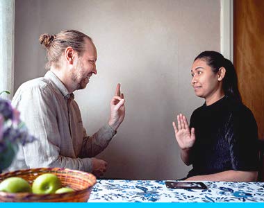 Two ASL signers are having a conversation, sitting by a kitchen table with a floral tablecloth and basket of apples on it.