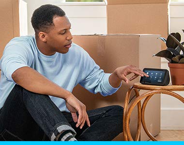 In a living room, next to moving boxes, a man sits on the floor and taps on the screen of an Echo Show device.