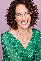 White woman smiling with curly red hair, big smile, bright green blouse and gold chains.