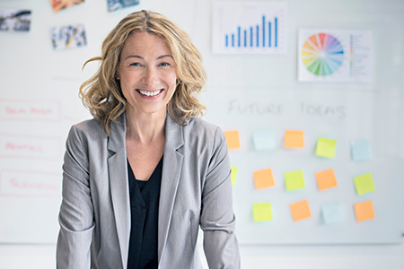 Woman wearing a suit, smiling in front of a wall with charts and sticky notes on it.