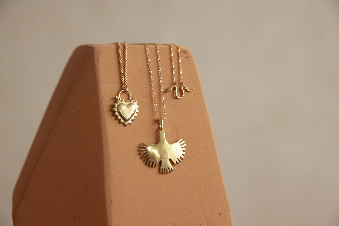 Three brass necklaces with heart, snake, and bird charms on pink wood block. Handmade jewelry in Oakland.