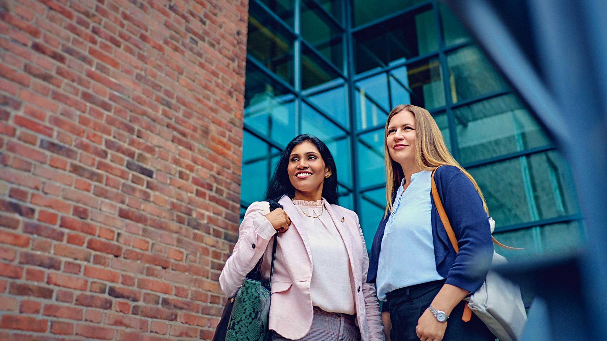 Two young women wearing pink and dark blue blazers and holding handbags standing together and smiling in front of a big building made of red bricks and big glass windows.