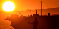 A silhouette of a man fishing off a jetty with a bright sun shining in the upper left corner