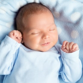 Newborn Baby Sleeping Smiling. Cute Infant Child in Wrap Bodysuit. New Born Little Boy smile in Blue Clothes lying on Blanket. Small Kid Face Close up Portrait holding Hands First